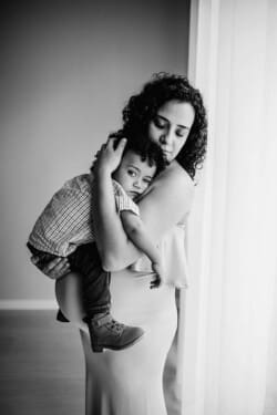 A pregnant mother holds her little boy close as they stand by a bright studio window
