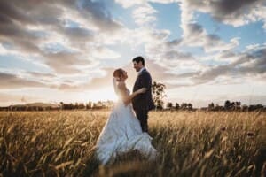 A groom holds his bride as they stand in a field of long grass with the sunsetting over Burnham Grove