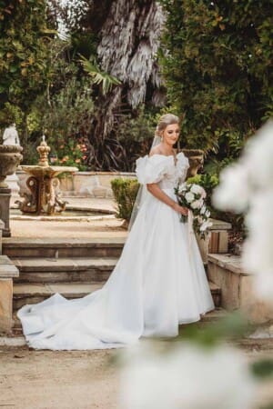 A bride stands in the gardens of Burnham Grove holding a white bouquet and wearing a flowing white dress