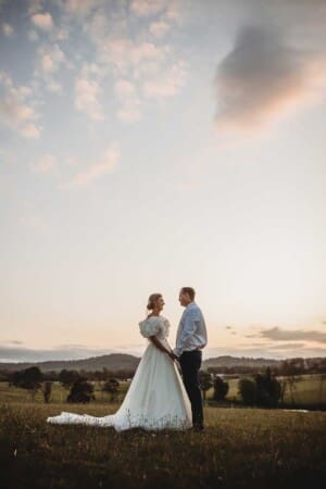 A bride and groom stand holding hands as the sun sets behind them over Burnham Grove