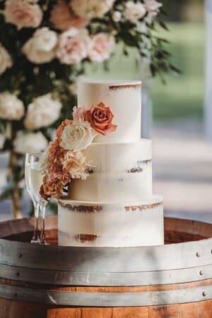 A wedding cake sits on a wine barrel with white icing covered in peach coloured roses