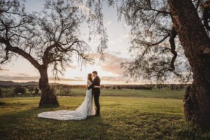 A couple stand beneath the Burnham Grove pepper trees with a golden sunset appearing behind them