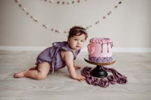 A baby girl in a purple jumpsuit crawls towards her pink birthday cake before smashing it