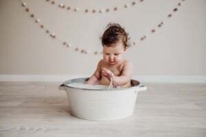 A baby girl sits in a white vintage tub and splashes water after her cake smash session