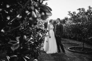 Black and white photograph of a bride and groom standing in a grove of trees at sunset