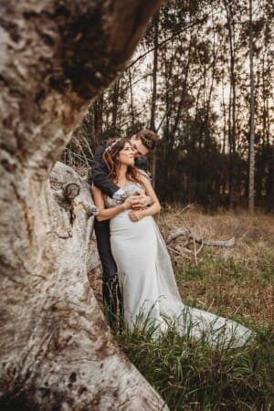 Newlyweds stand against a huge tree trunk in a field at Gledswood after their wedding day