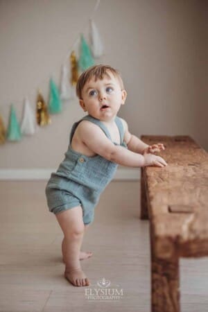 A baby balances himself against a wooden bench while standing in a bright studio