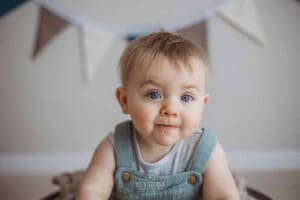 A baby boy sits in a bright studio wearing a green romper with bunting behind him