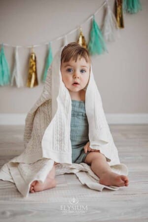 A baby boy sits under a white blanket on a white wood studio floor with bunting behind him