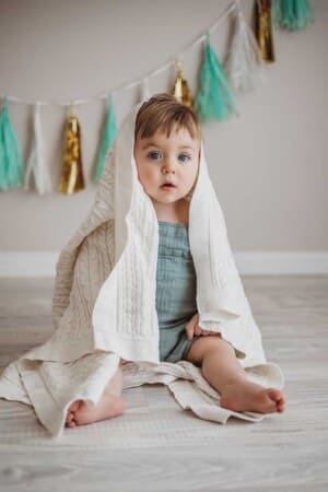 A baby boy wearing a green romper sits under a white knitted blanket in a bright neutral studio