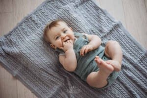 A smiley baby lays on his back on a grey blanket in a brightly lit studio