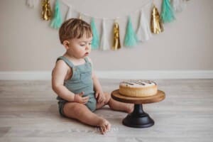 A baby boy sits beside a cake stand with a first birthday cake in a studio