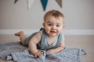A baby boy in a soft green romper lays on his tummy on a grey blanket