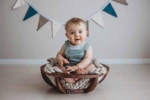 A baby boy sits smiling in a large wooden bowl with coloured bunting behind him