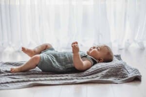 A baby lays on his back on a grey blanket in front of a large bright studio window