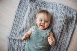 A baby boy in a soft green romper lays on his back on a grey blanket beside a window