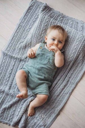 A baby boy sucks his thumb as he lays on his back on a grey blanket