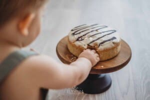 A first birthday cake sits on a wooden cake stand as a baby's hand grabs at it