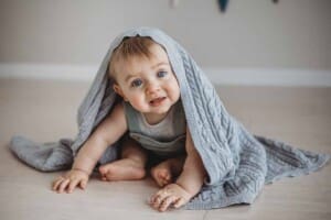 A baby boy smiles as he peeks out from under a grey blanket
