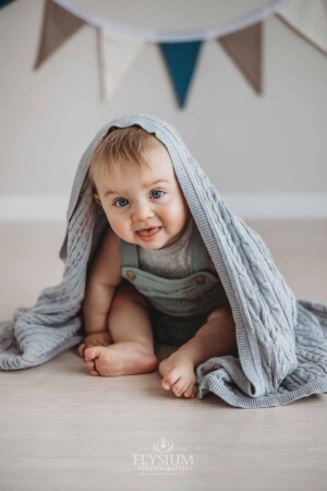 A baby boy plays peek-a-boo sitting on a floor under a grey blanket