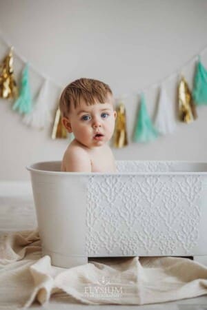 A baby boy sits in a white vintage bath tub after his cake smash session