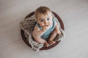 A baby boy sits on a beige bump blanket in a wooden bowl looking up at the camera
