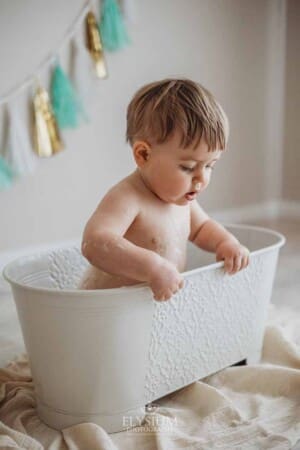 A little baby leans over the edge of a small bathtub in a studio