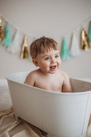 A baby boy smiles as he sits in a white vintage bathtub after his cake smash session