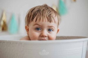 A baby with blue eyes peers over the edge of a white bathtub