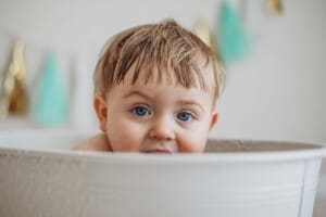 A baby boy with blue eyes gazes over the edge of a white bathtub after his cake smash session