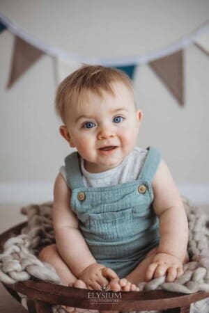 A baby boy wearing a green romper sits in a large wood bowl with bunting hanging behind him