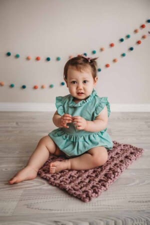 A baby girl in a green romper sits on a pink bump blanket for her first birthday milestone session