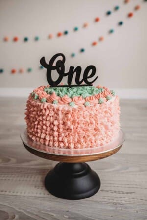 A peach and mint coloured frosted cake with a ONE cake topper sits on a wood and metal cake stand in a studio