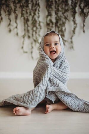 A baby boy smiles as he wraps a grey knitted blanket around himself as he sits in a bright studio