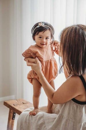 A baby girl holds hands with her mum and stands in her lap during her milestone session