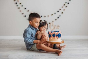 Siblings sit on a white wood floor next to a colourful cake before smashing it