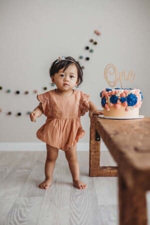 A baby girl stands beside a colourful cake sitting on a wooden bench