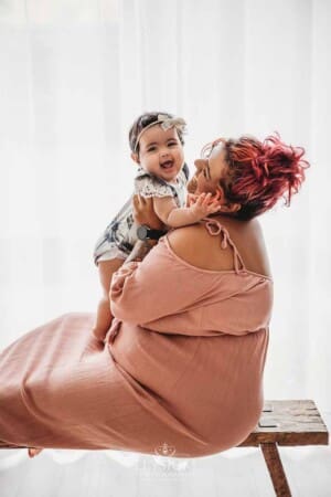 A baby girl sitting in her mum's lap on a timber bench next to a bright studio window