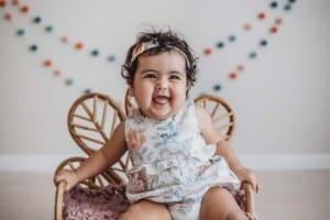 A smiley baby girl sits on a little chair in a floral playsuit during her studio session