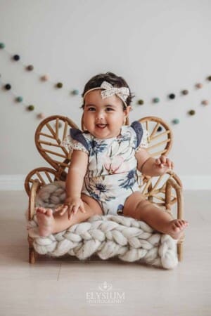 A baby girl in a floral romper sits in a studio prop for a sitter session