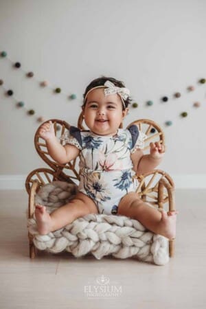 A little girl smiles as she sits in a tiny rattan chair for her baby photo session in a bright studio