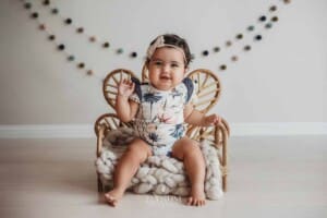 A baby wearing a floral romper sits in a studio prop chair and smiles for a photo
