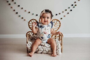 A baby sitting on a tiny rattan chair smiles and enjoys a milestone session