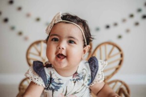 A baby dressed in a floral romper and bow headband smiles sitting in a studio prop