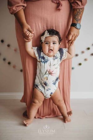 A mother wearing a pink dress hold her baby's hands as she stands in front wearing a floral romper
