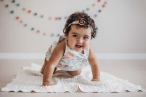 A crawling baby smiles as she sits on a white blanket wearing a floral romper
