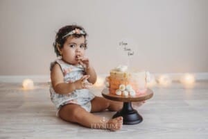 A baby girl in a floral romper sits beside a birthday cake that sits on a cake stand