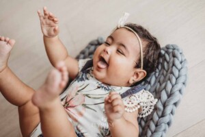 A baby girl lays on a grey bump blanket and smiles as she grabs her feet