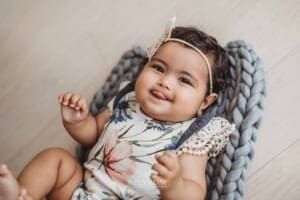 A baby girl lays on her back on a grey bump blanket and sticks out her tongue as she smiles