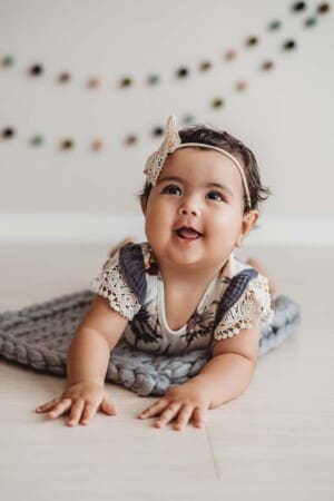 A baby lays on a knitted grey bump blanket and smiles during her studio sitter session